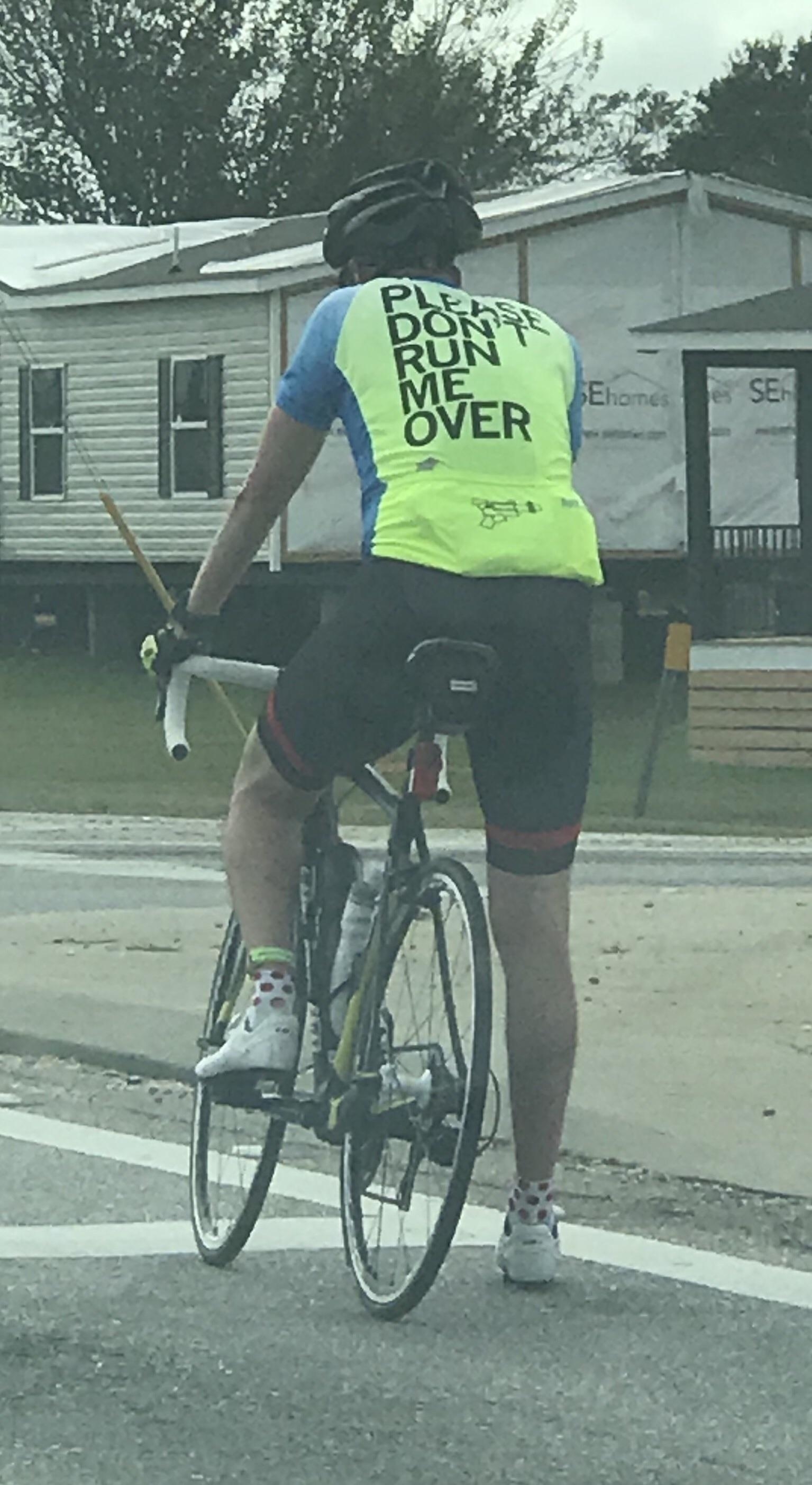 Cyclist on a road bike wearing a shirt that reads "Please Don't Run Me Over" rides near mobile homes