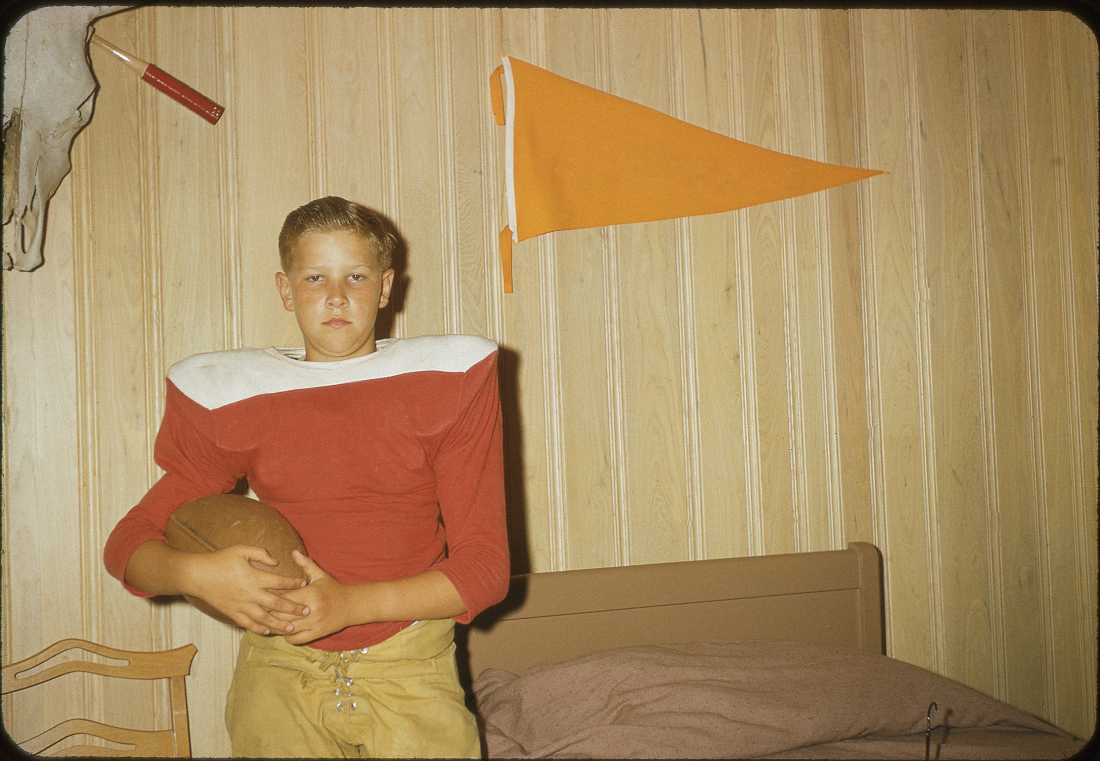 A young boy in a football uniform stands in a room holding a football, with a bed and an orange pennant on the wooden paneled wall behind him
