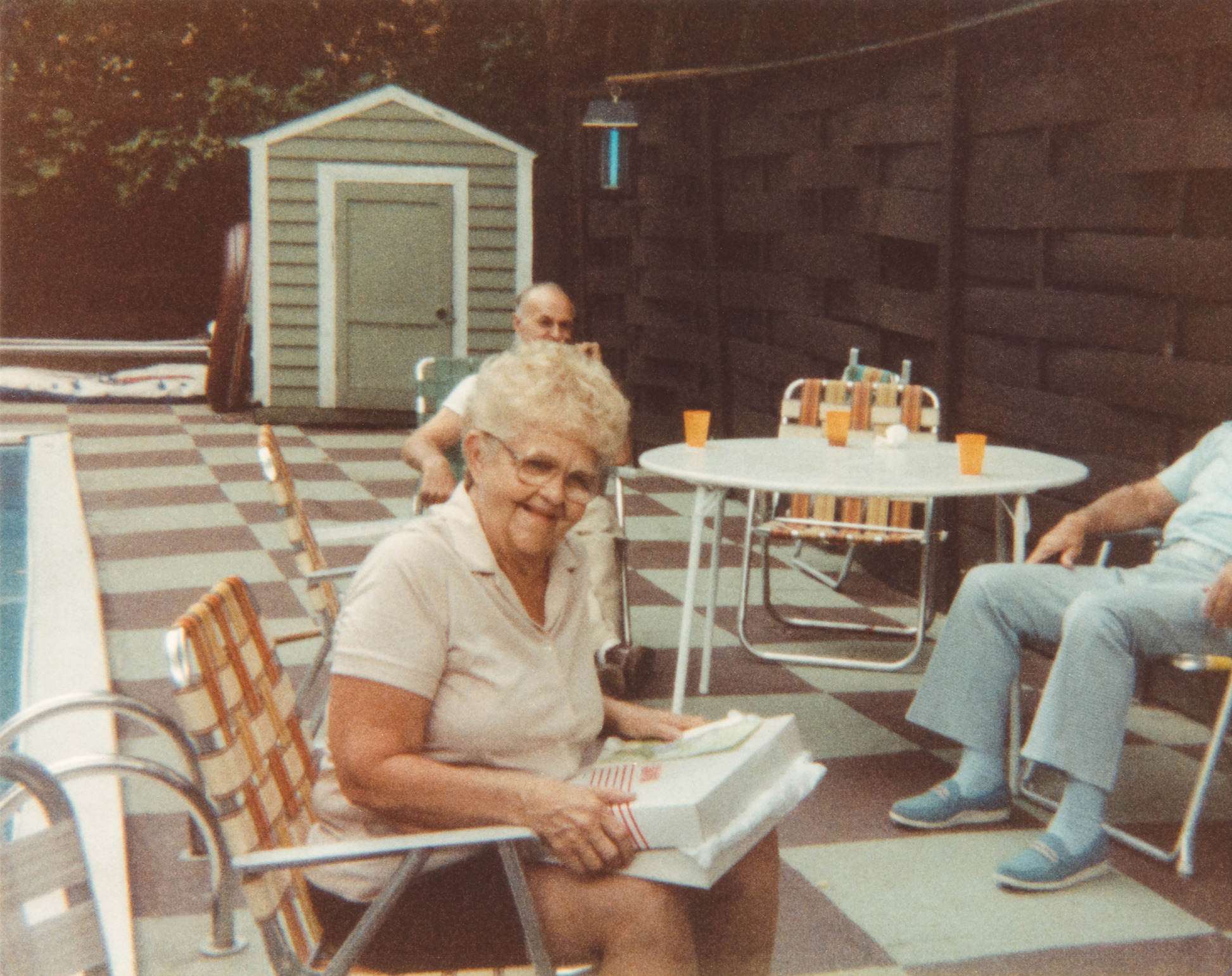 Three elderly individuals sit by a pool, with one woman smiling at the camera while holding a wrapped gift. A shed and table with drinks are in the background. Names unknown