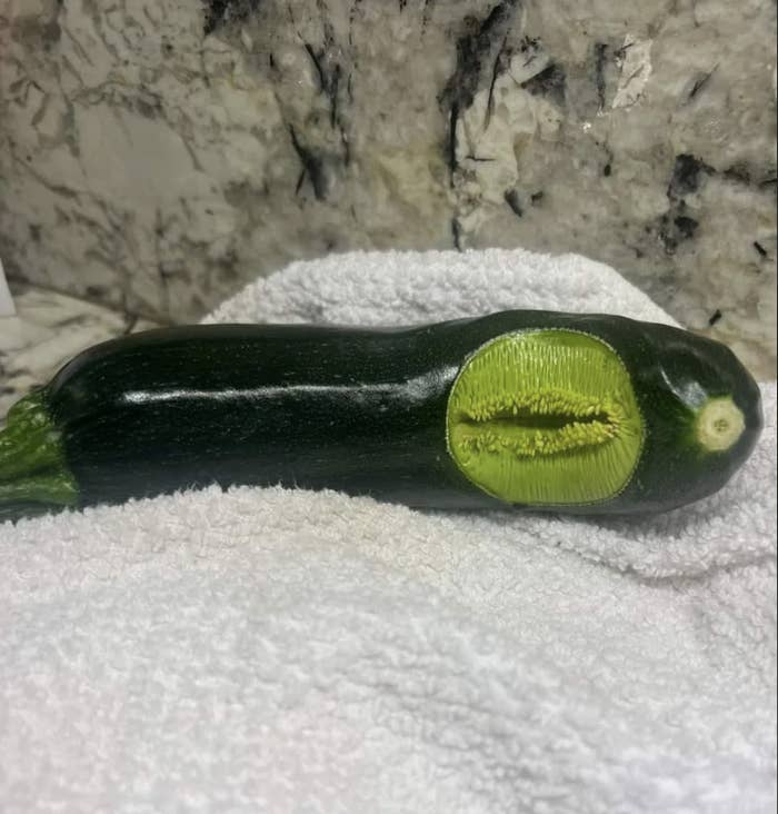 A zucchini with a section of green embroidery on its side rests on a white towel, set against a marbled background. There are no people in the image