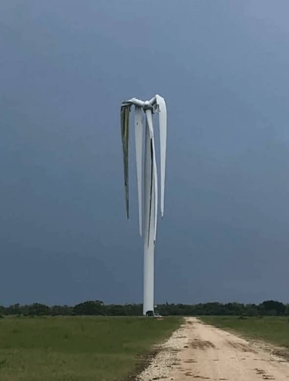 A damaged wind turbine stands alone in a grassy field with bent and twisted blades, under a dark cloudy sky. A dirt path leads towards it