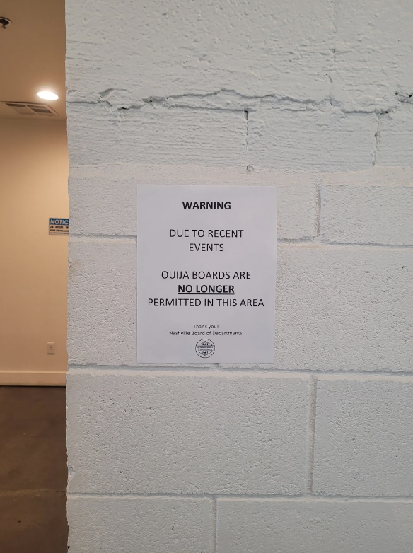A sign on a white brick wall reads: "WARNING. Due to recent events, Ouija boards are no longer permitted in this area. Thank you! Nashville Board of Dreamerevents."