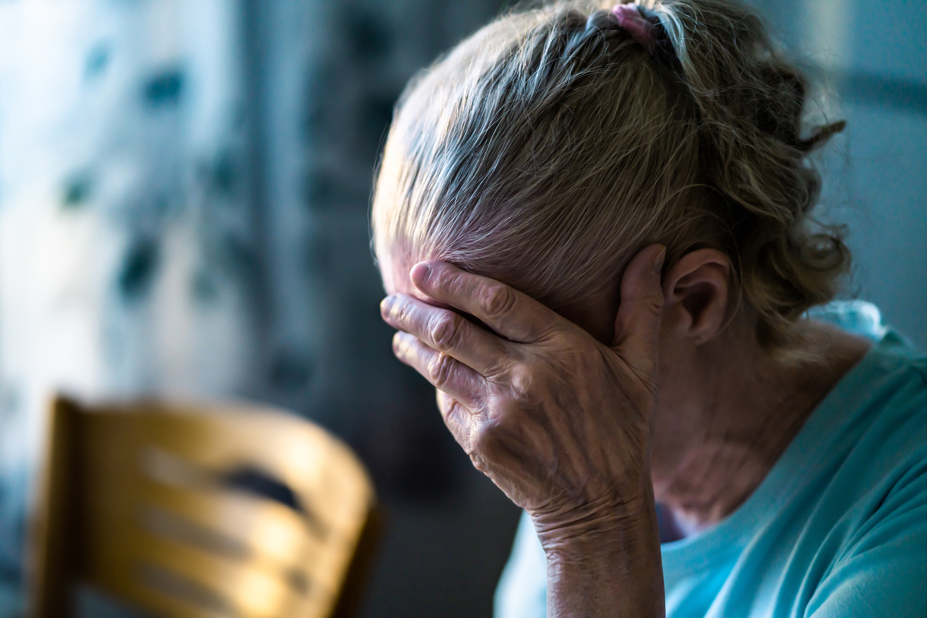 Elderly woman, hands covering face, appears distressed while seated in a room with a chair and blurred background