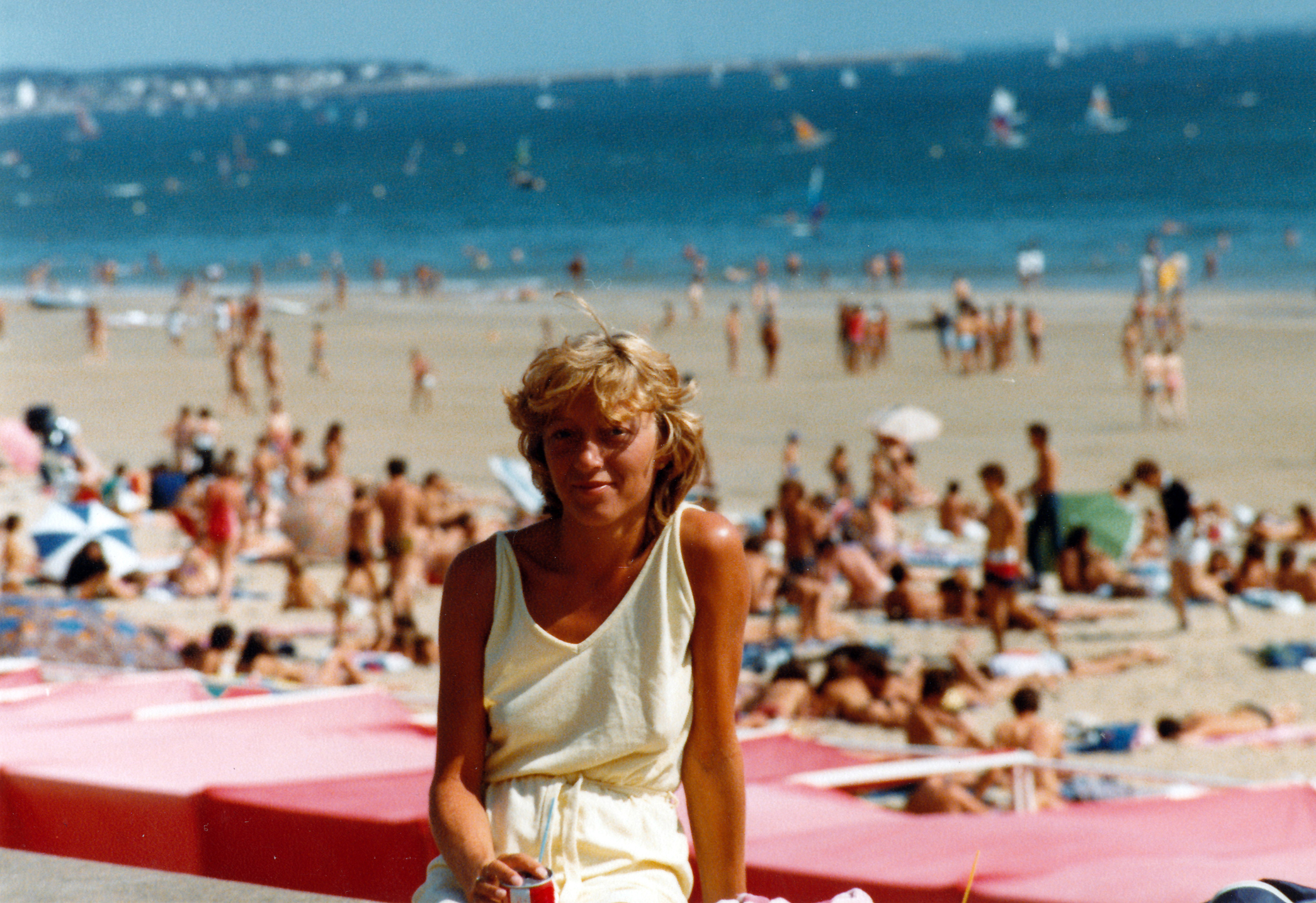 A person in light summer clothes sits and smiles on a busy beach with many people sunbathing and swimming in the background