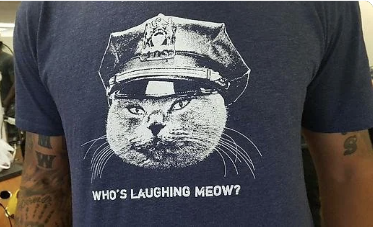Person wearing t-shirt featuring a cat with a police hat and text "WHO'S LAUGHING MEOW?"