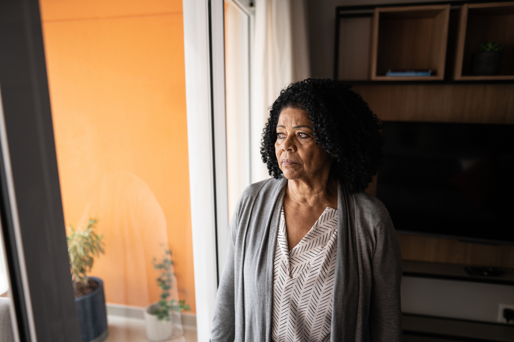 An older woman with curly hair gazes out of a window, wearing a patterned blouse and a light cardigan, standing in a modern living room with a TV and potted plants