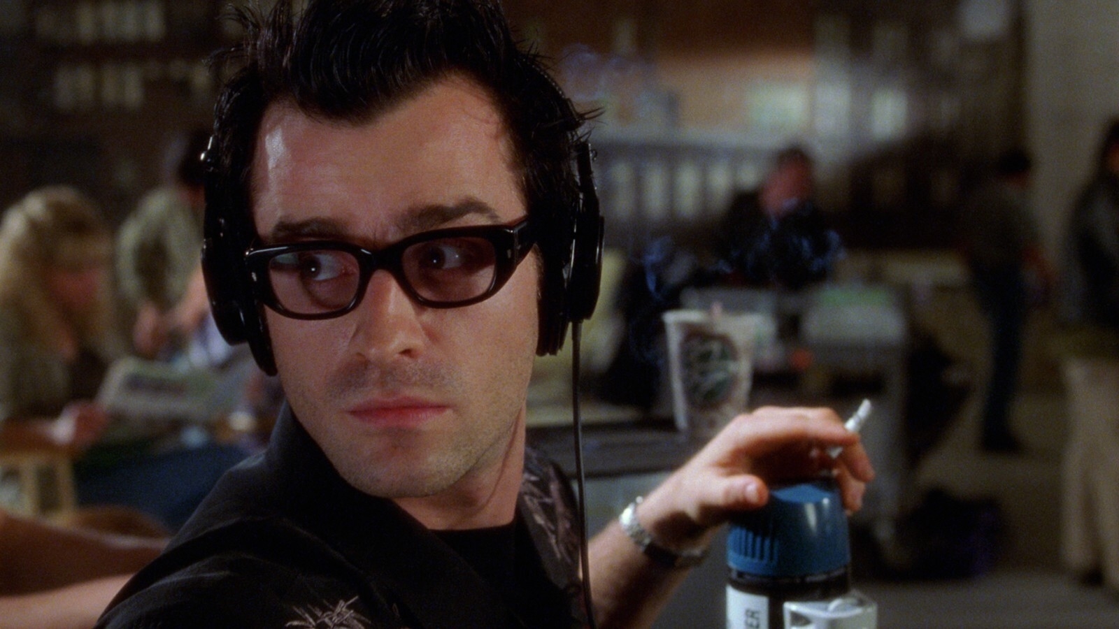 A man with dark hair, wearing black-rimmed glasses and headphones, holds a blue and white object while looking to his left in a busy indoor setting