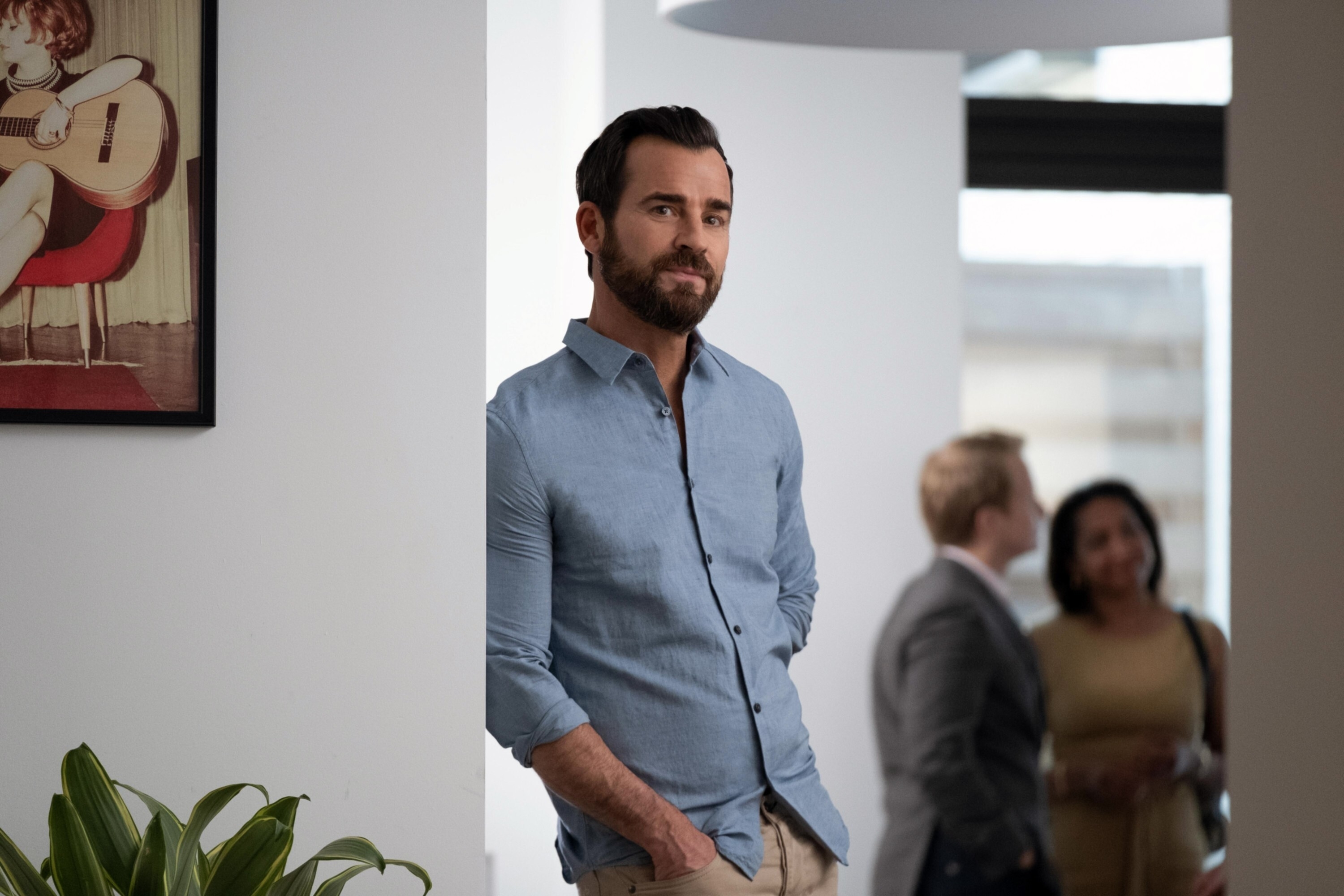 Justin Theroux in a button-up shirt, leaning against a wall with two people blurred nearby in "False Positive"