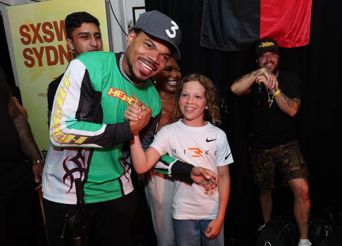 Chance the Rapper wearing a green and black track jacket, smiling and shaking hands with a young boy in a white shirt at an event with several people in the background