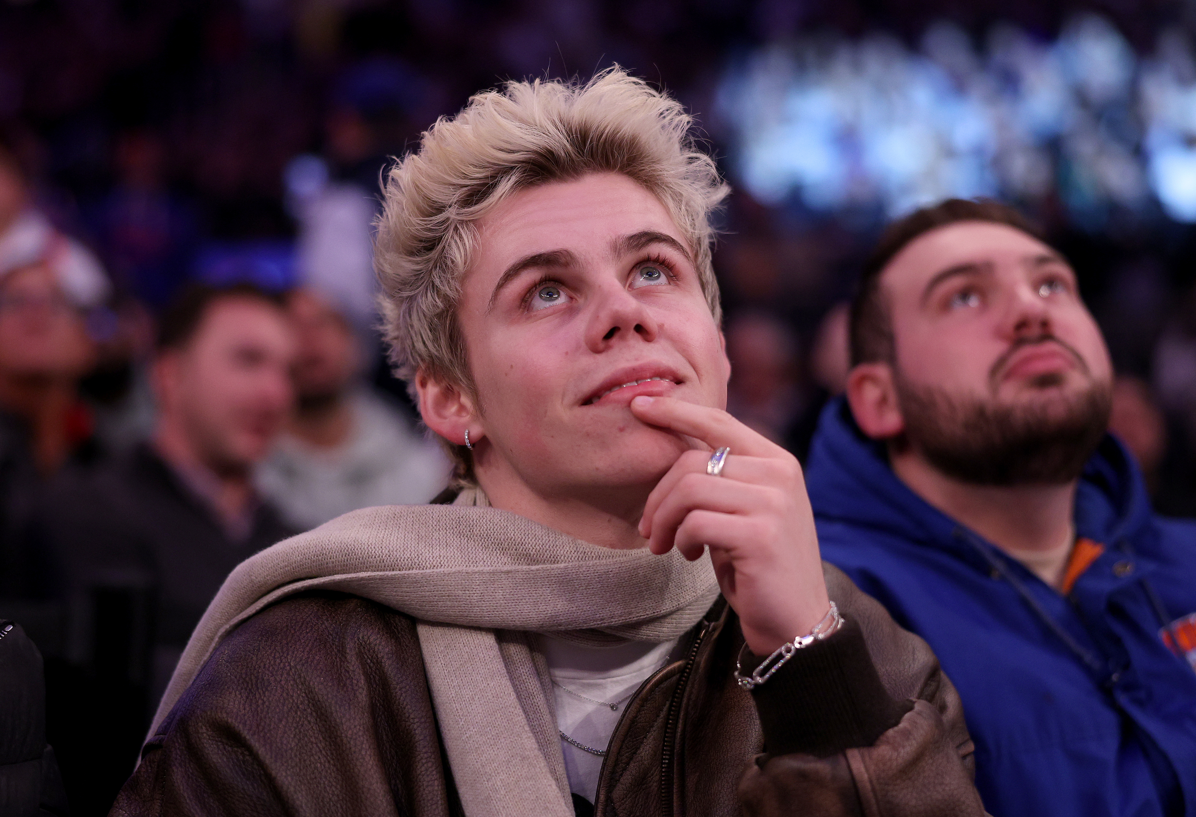 The Kid Laroi wearing a scarf and a jacket, sitting next to a bearded man, looking thoughtful at a sports event