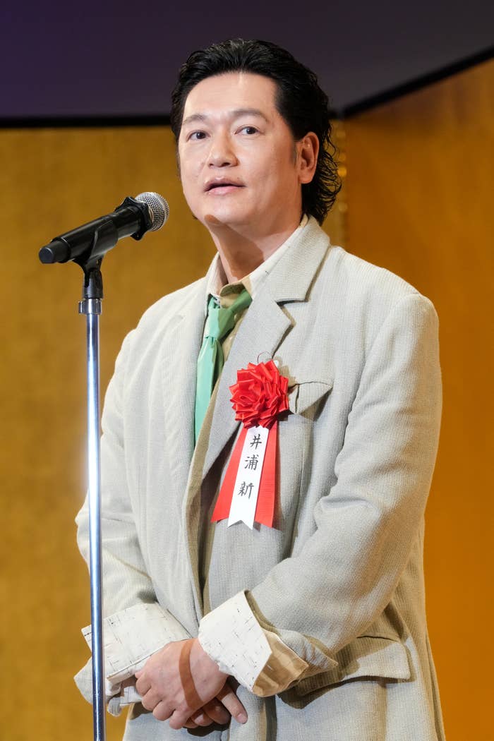 井浦新がスピーチ中、大きな花のコサージュを付けたベージュのスーツを着ています。