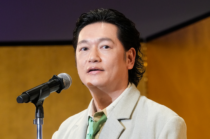 井浦新がスピーチ中、大きな花のコサージュを付けたベージュのスーツを着ています。