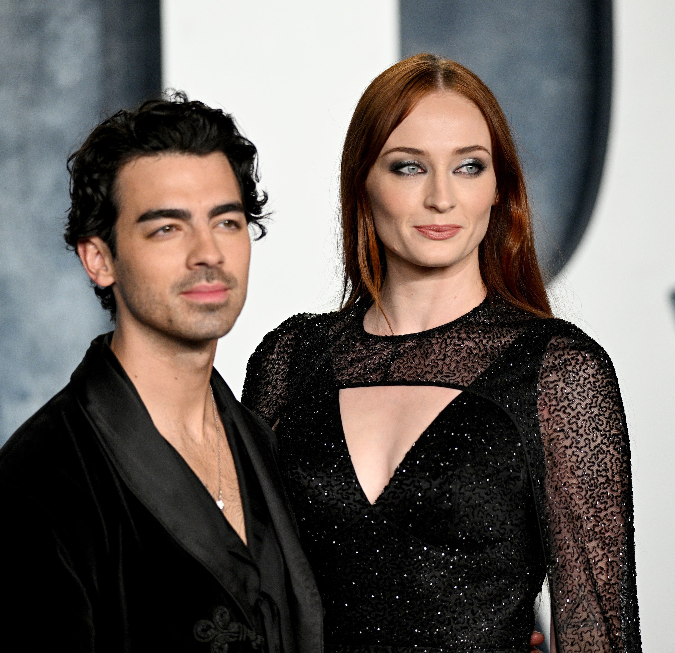 Joe Jonas and Sophie Turner stand together on a red carpet, with Joe in a black outfit and Sophie wearing a black dress with sheer sleeves