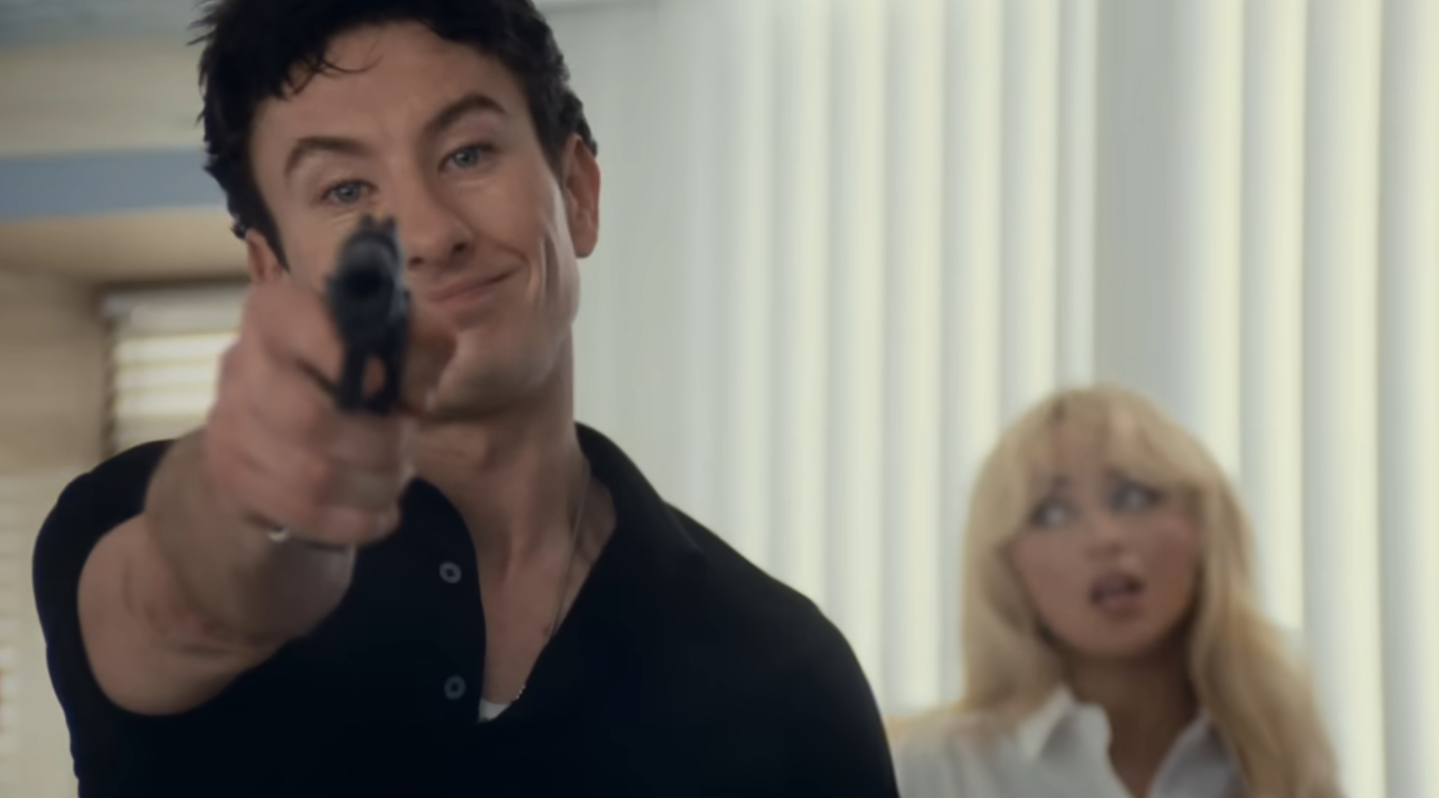 Barry Keoghan, in the Please Please Please music video, points a gun with a confident smirk, while Sabrina Carpenter stands in the background with a surprised expression