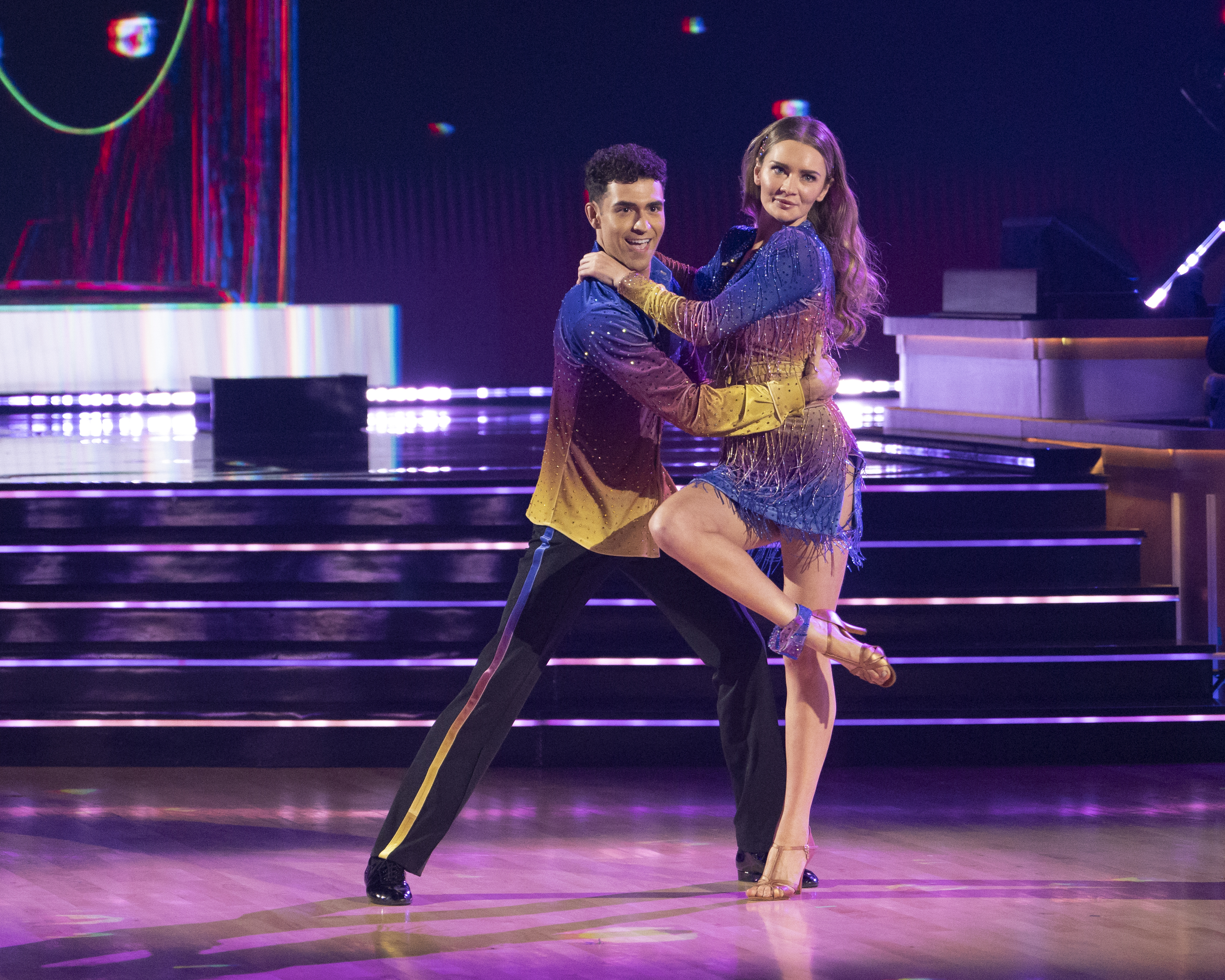 Anna and Ezra perform a ballroom dance on stage, wearing matching dazzling outfits