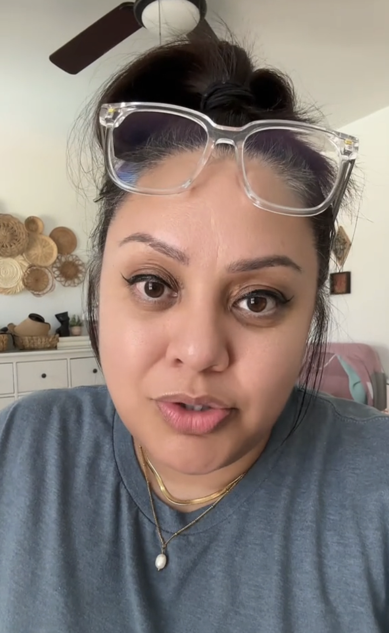A person with glasses on their head, wearing a casual shirt and a necklace, talking to the camera in a room with decorative baskets on the wall