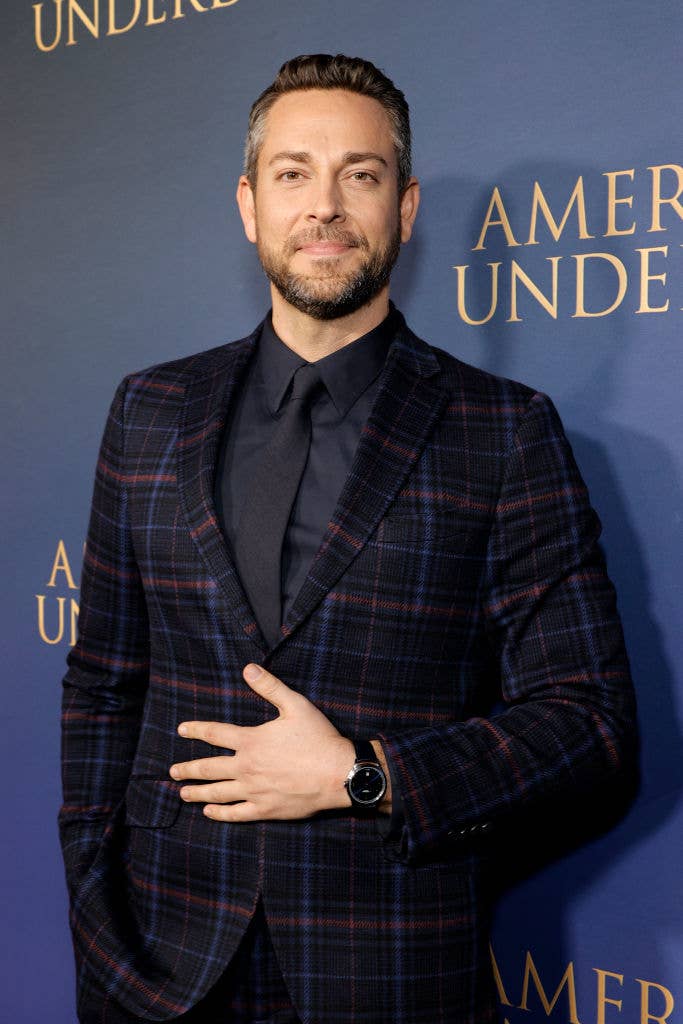 Zachary Levi poses on the red carpet, wearing a dark plaid suit with a black shirt and tie, against a backdrop with the text "AMER UND."