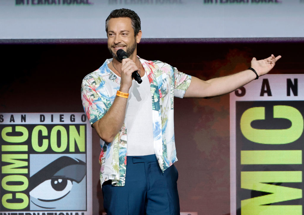 Zachary Levi speaks on stage at San Diego Comic-Con, wearing a floral shirt, white T-shirt, and blue pants