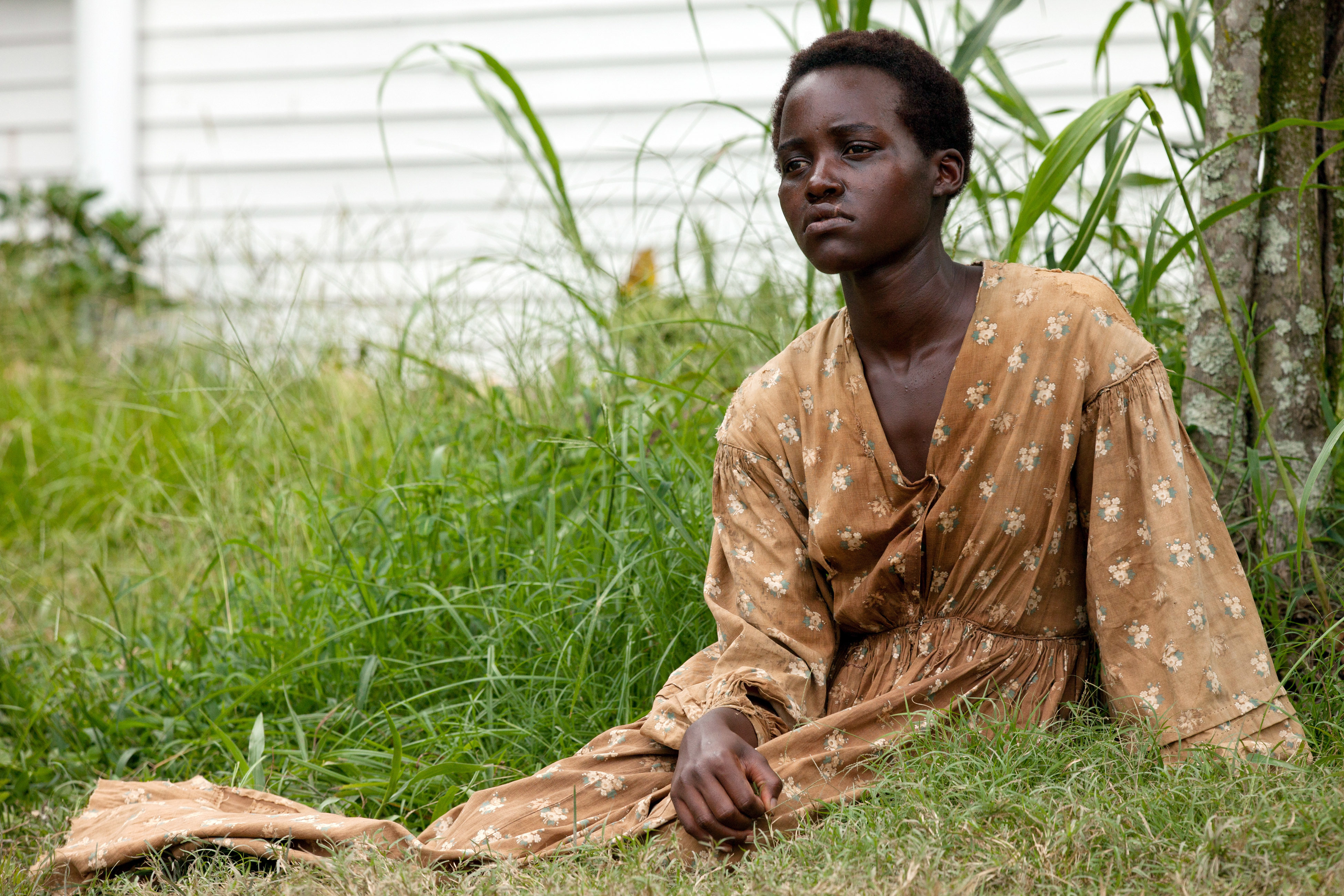 Lupita Nyong'o in a patterned dress, sitting on grass, appears thoughtful in front of a house with tall plants around her