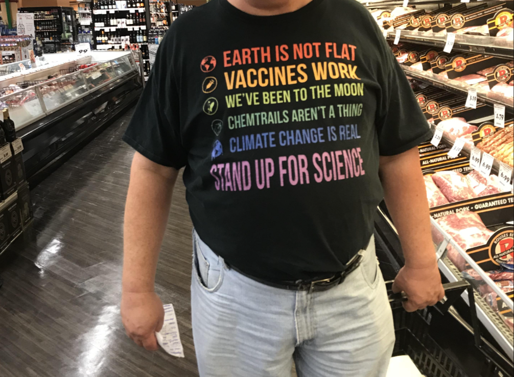Person wearing a t-shirt in a grocery store. The t-shirt reads: "Earth is not flat, vaccines work, we've been to the moon, chemtrails aren't a thing, climate change is real, stand up for science."