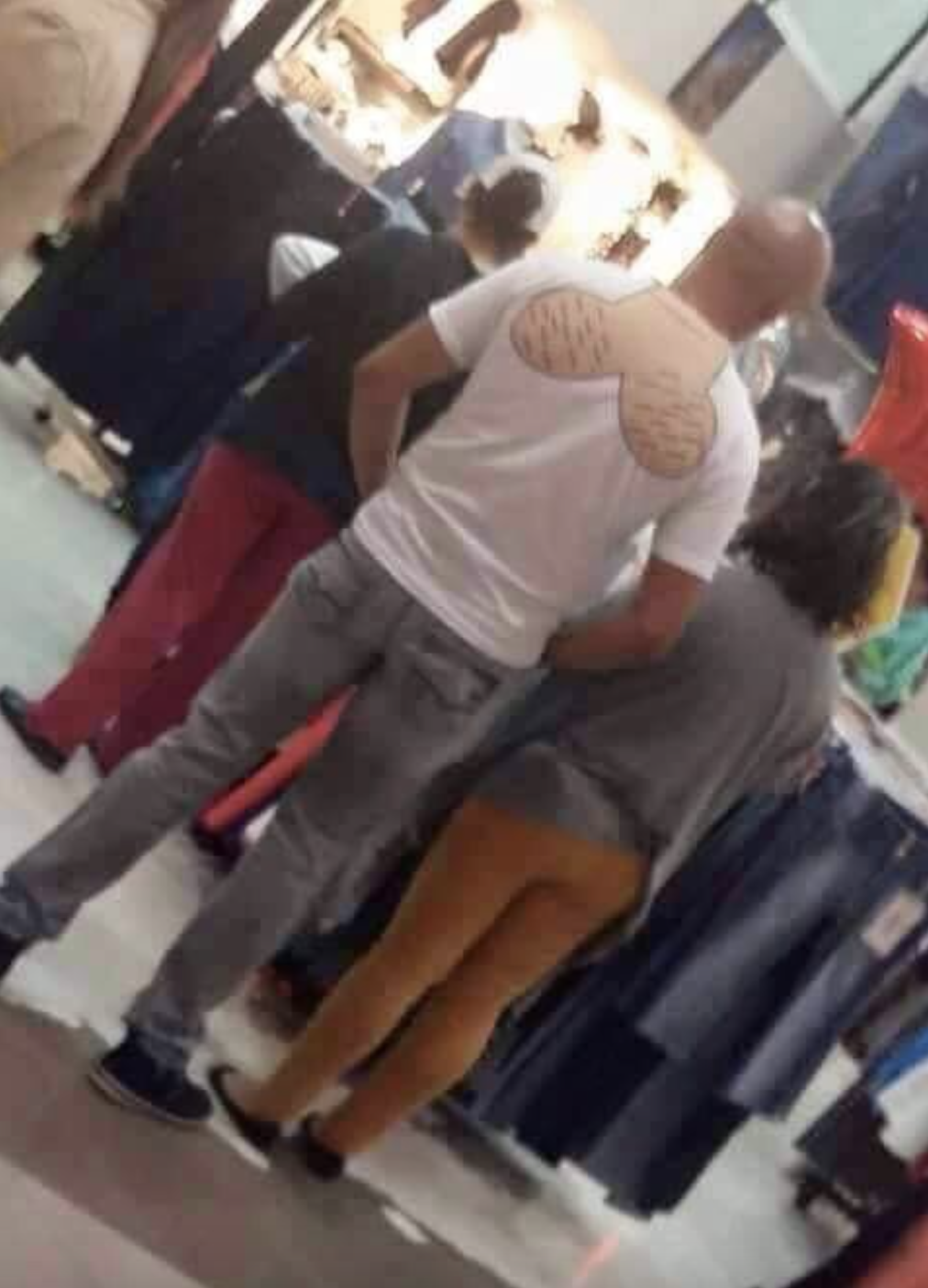 A man with a humorous shirt showing muscles on the back stands next to a woman in a store. They appear to be shopping
