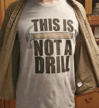 Person wearing a plaid shirt open to reveal a t-shirt with a hammer graphic and text reading "THIS IS NOT A DRILL."