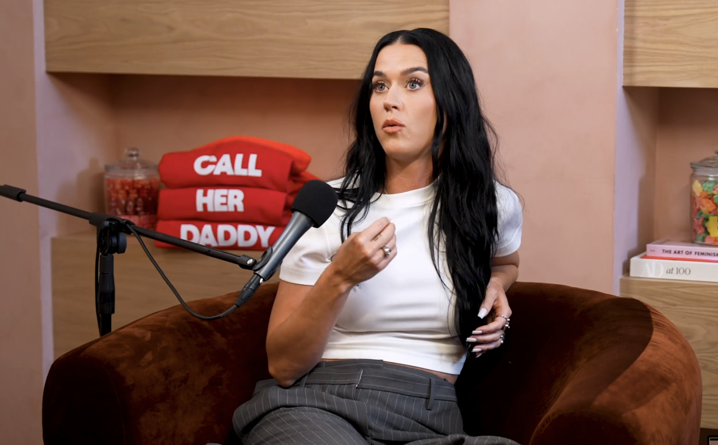 Katy Perry, in a white crop top and grey pants, sits in a podcast studio with a microphone, shelves with books, and a &quot;Call Her Daddy&quot; sign behind her