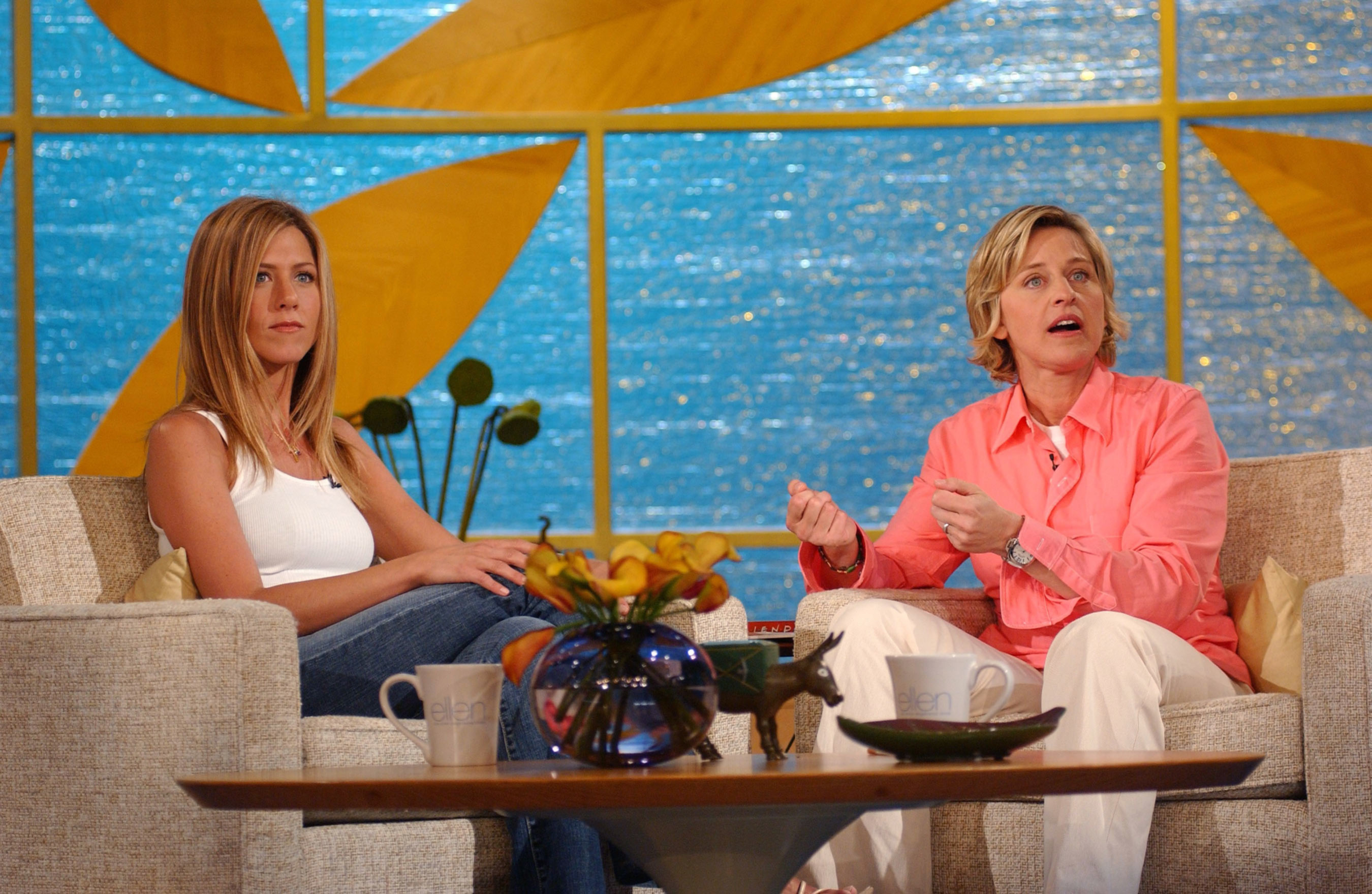 Jennifer Aniston and Ellen DeGeneres sit on a talk show set. Aniston is wearing a white top and jeans, while DeGeneres wears a pink shirt and light pants