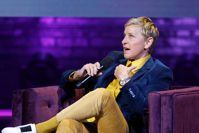 Ellen DeGeneres is seated, holding a microphone, gesturing while speaking. She is wearing a blazer, yellow shirt, and yellow pants