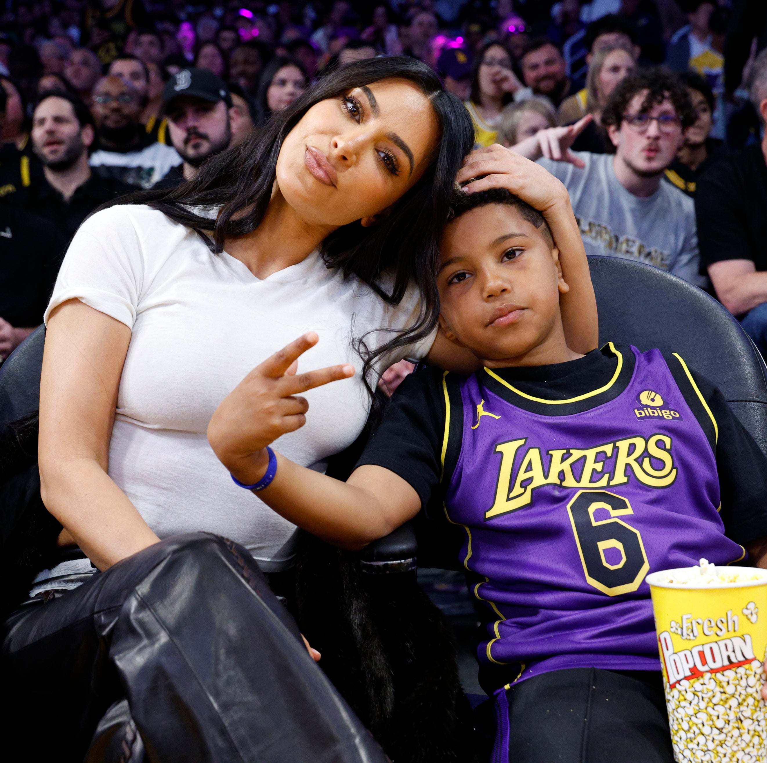 Kim Kardashian sits courtside with her son Saint West at a Lakers game. Saint is holding a cup of popcorn and wearing a Lakers jersey