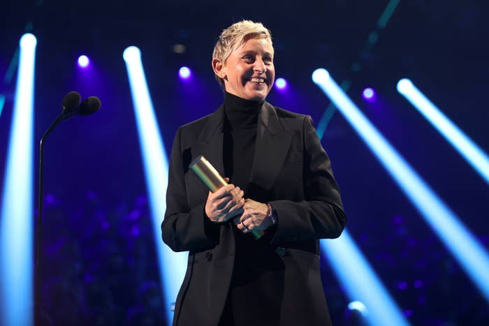 Ellen DeGeneres stands on stage holding an award, wearing a suit and turtleneck, smiling, and in front of colorful stage lights