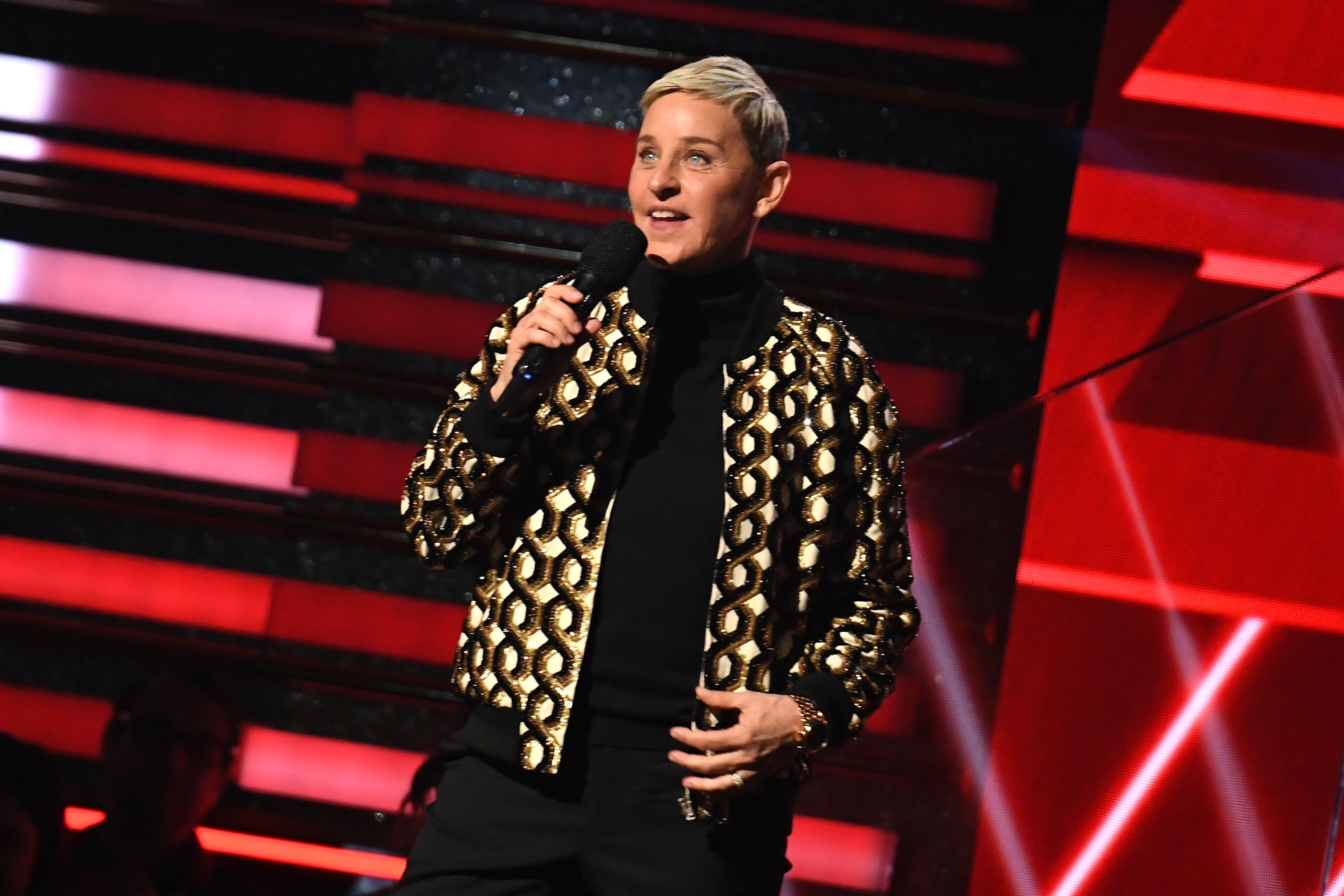 Ellen DeGeneres speaks on stage wearing a gold-patterned jacket, holding a microphone