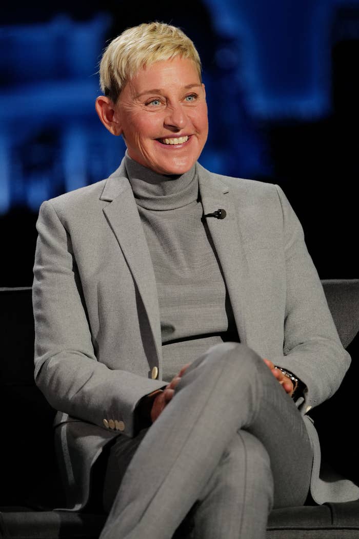 Ellen DeGeneres smiling while seated and wearing a suit with a turtleneck on a talk show set