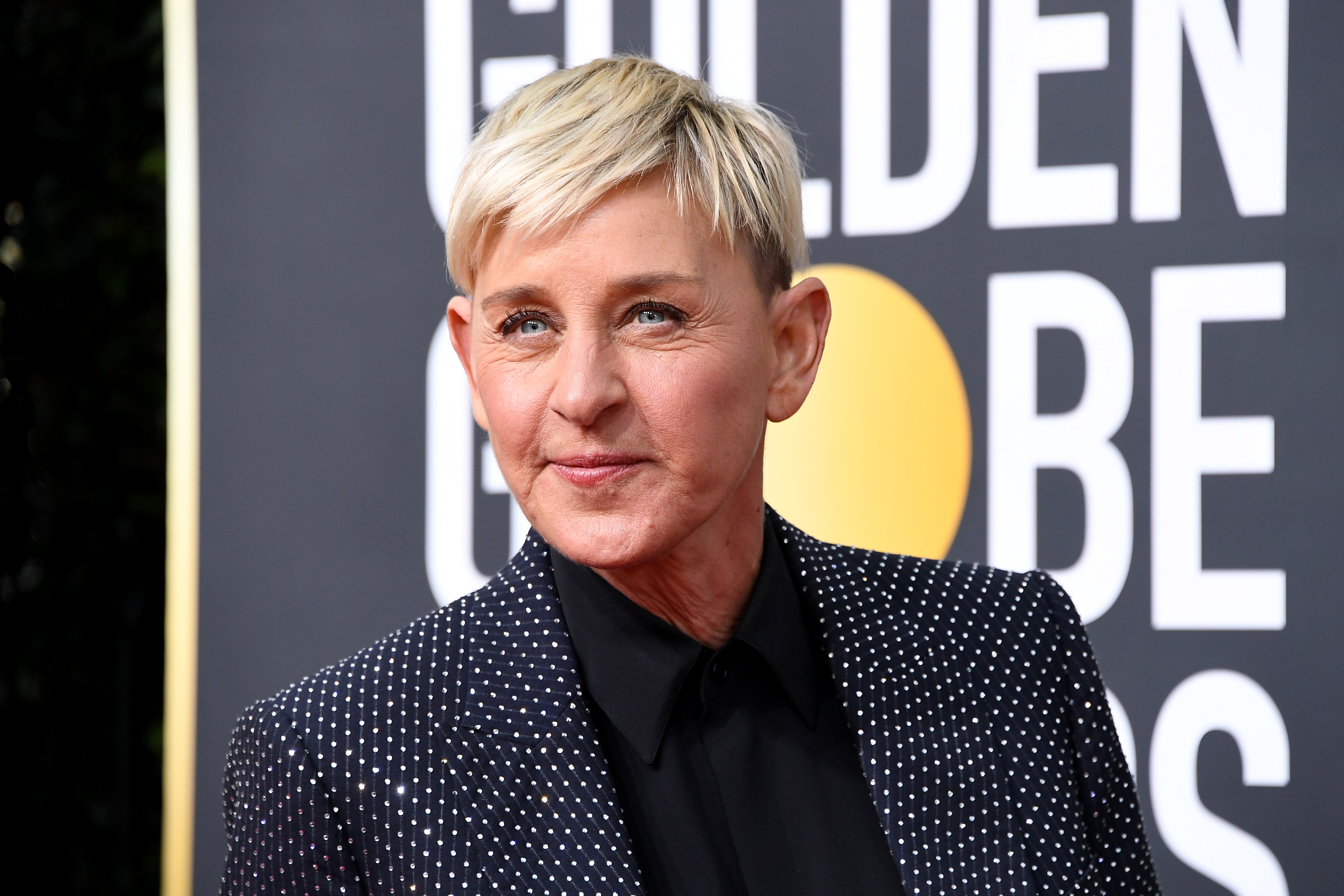 Ellen DeGeneres at an event, wearing a patterned suit jacket over a black shirt, with a backdrop featuring the word "GOLDEN."