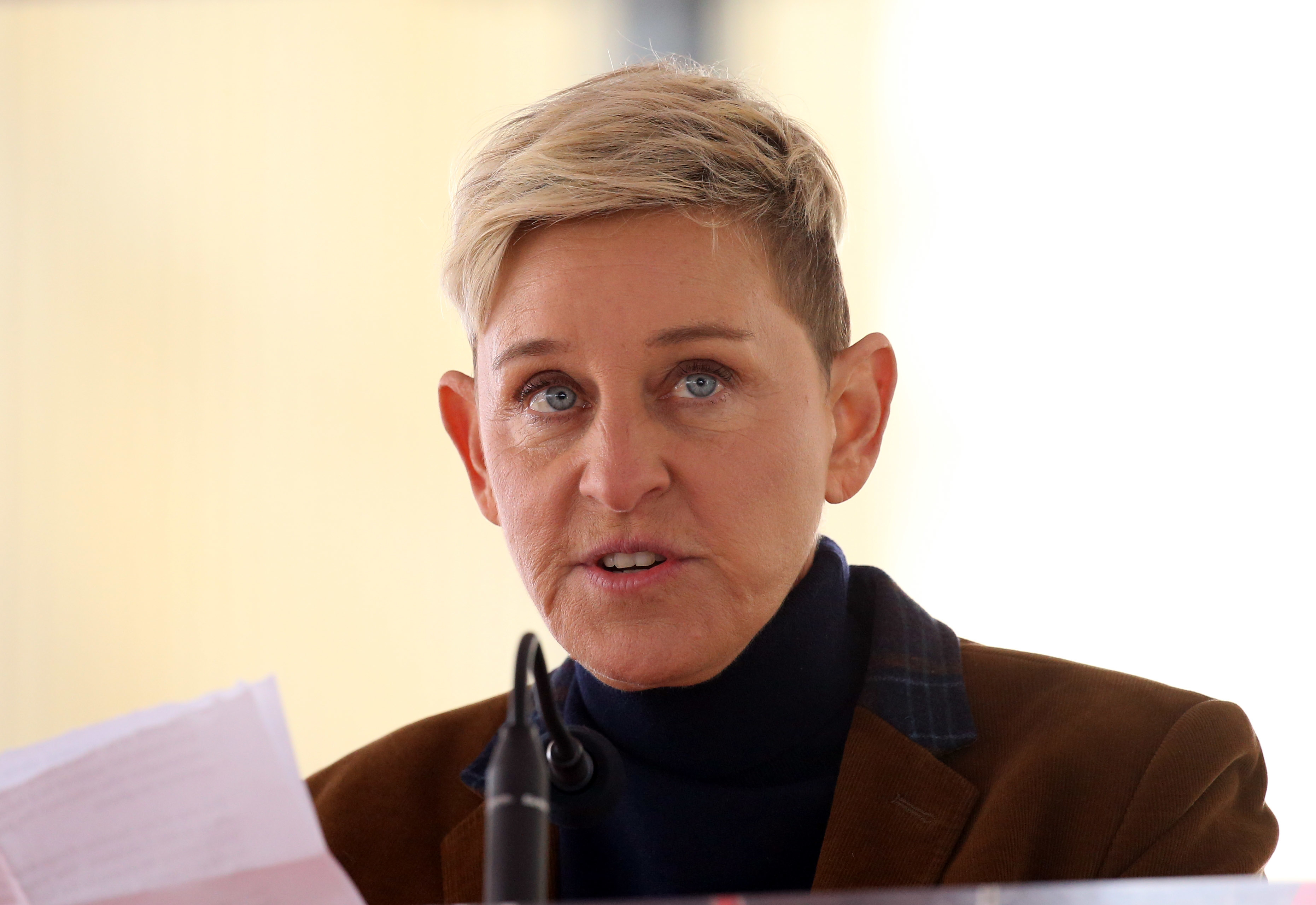 Ellen DeGeneres speaks at a podium, holding a piece of paper. She is wearing a black turtleneck and a brown jacket