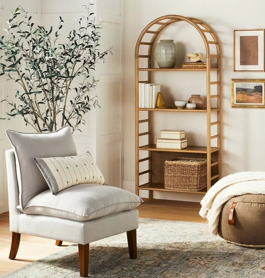 A cozy living room corner with a cushioned chair, a pillow, a pouf, and a wooden arch bookshelf filled with decor items