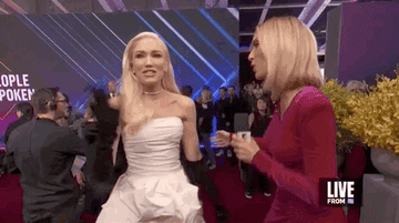 Gwen Stefani wears a white dress and black gloves at an event, talking with a reporter in a magenta dress. &quot;LIVE FROM&quot; is visible on the corner of the screen