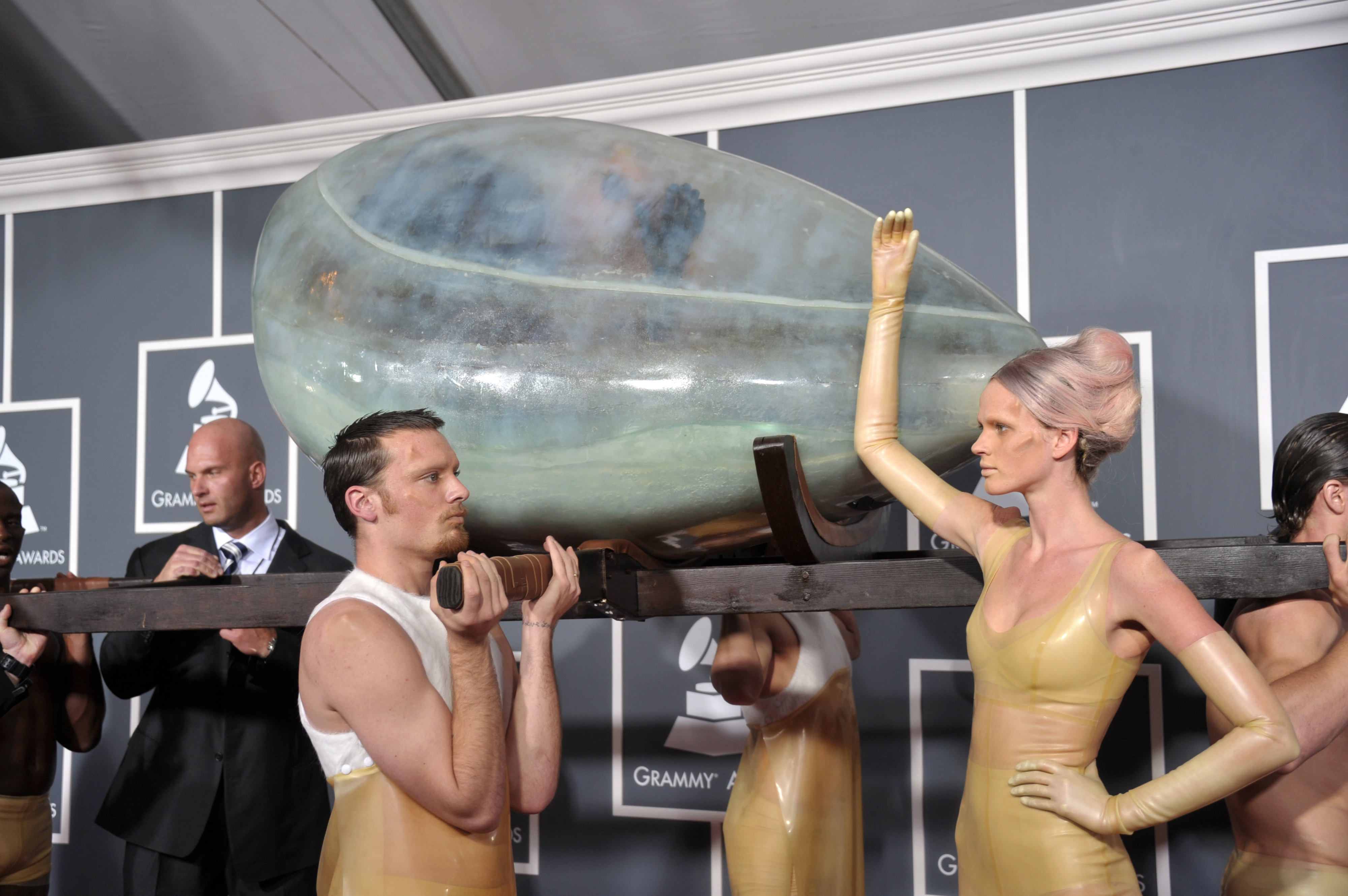 Lady Gaga being carried in an egg-like vessel by several people in latex bodysuits at the Grammy Awards red carpet