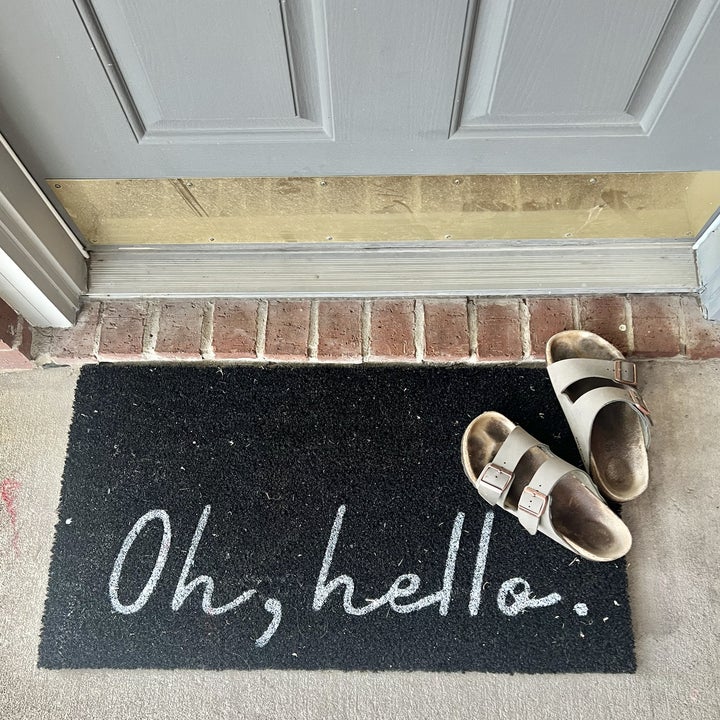 A black doormat with "Oh, hello.." written on it is placed outside a door with a pair of brown sandals beside it
