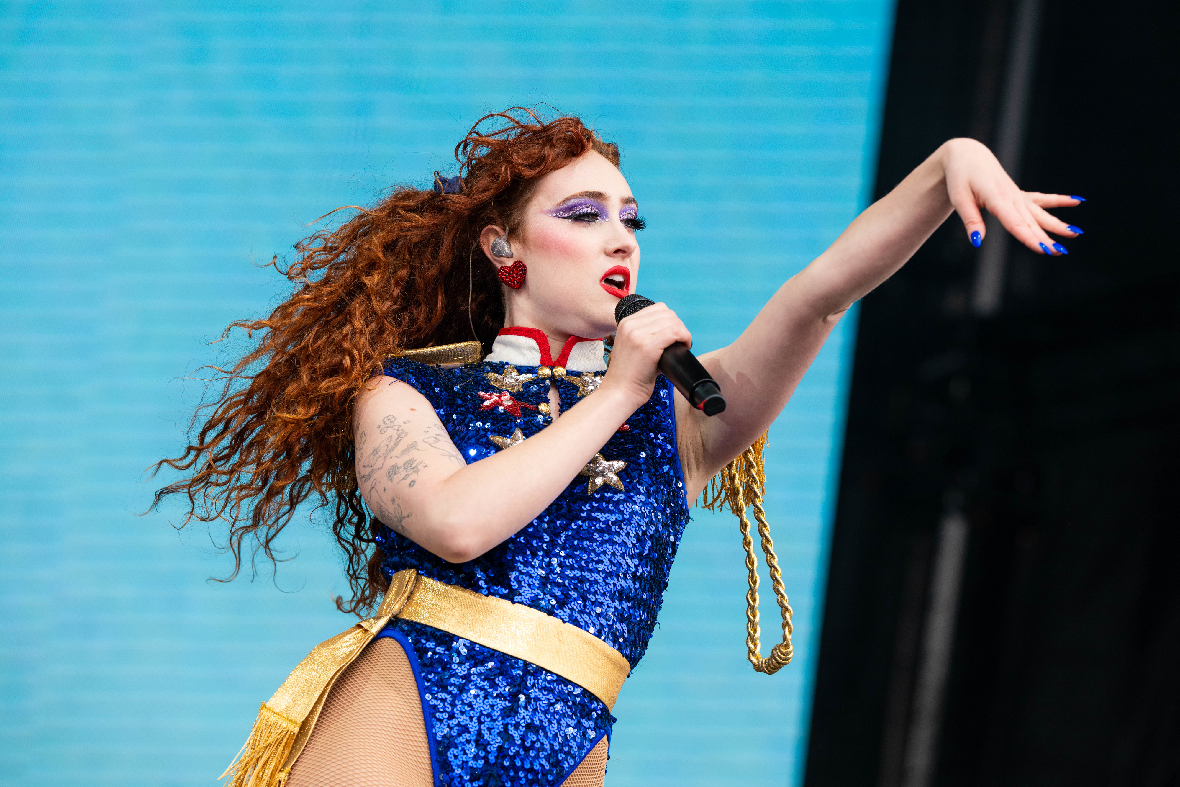 Chappell Roan with long curly hair on stage, wearing a sparkling blue bodysuit with gold accents, holding a microphone mid-performance