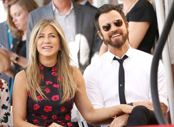 Jennifer Aniston in a floral dress and Justin Theroux in a shirt and dark tie, smiling while seated at an event