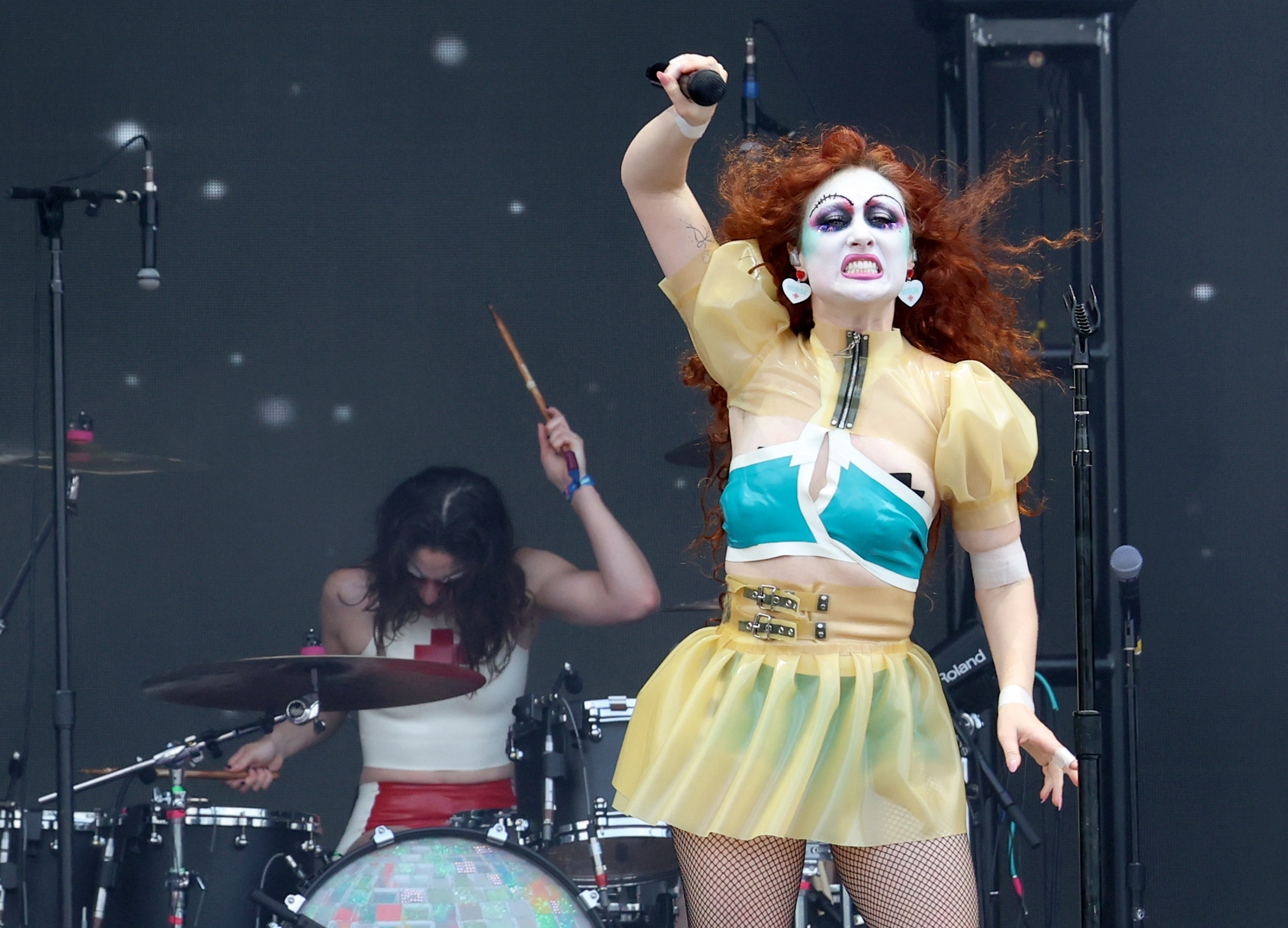 Chappell Roan on stage wearing a bold outfit with a green top and yellow skirt, and dramatic makeup