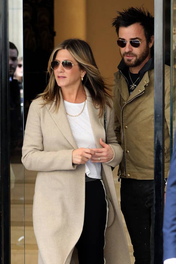 Jennifer Aniston and Justin Theroux stepping out together. Jennifer wears a beige coat, and Justin wears a khaki jacket and sunglasses