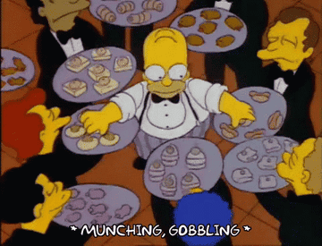 Homer Simpson from The Simpsons is dressed as a waiter, surrounded by people offering plates of food, while he reaches out eagerly. Text reads, &quot;*MUNCHING, GOBBLING*&quot;