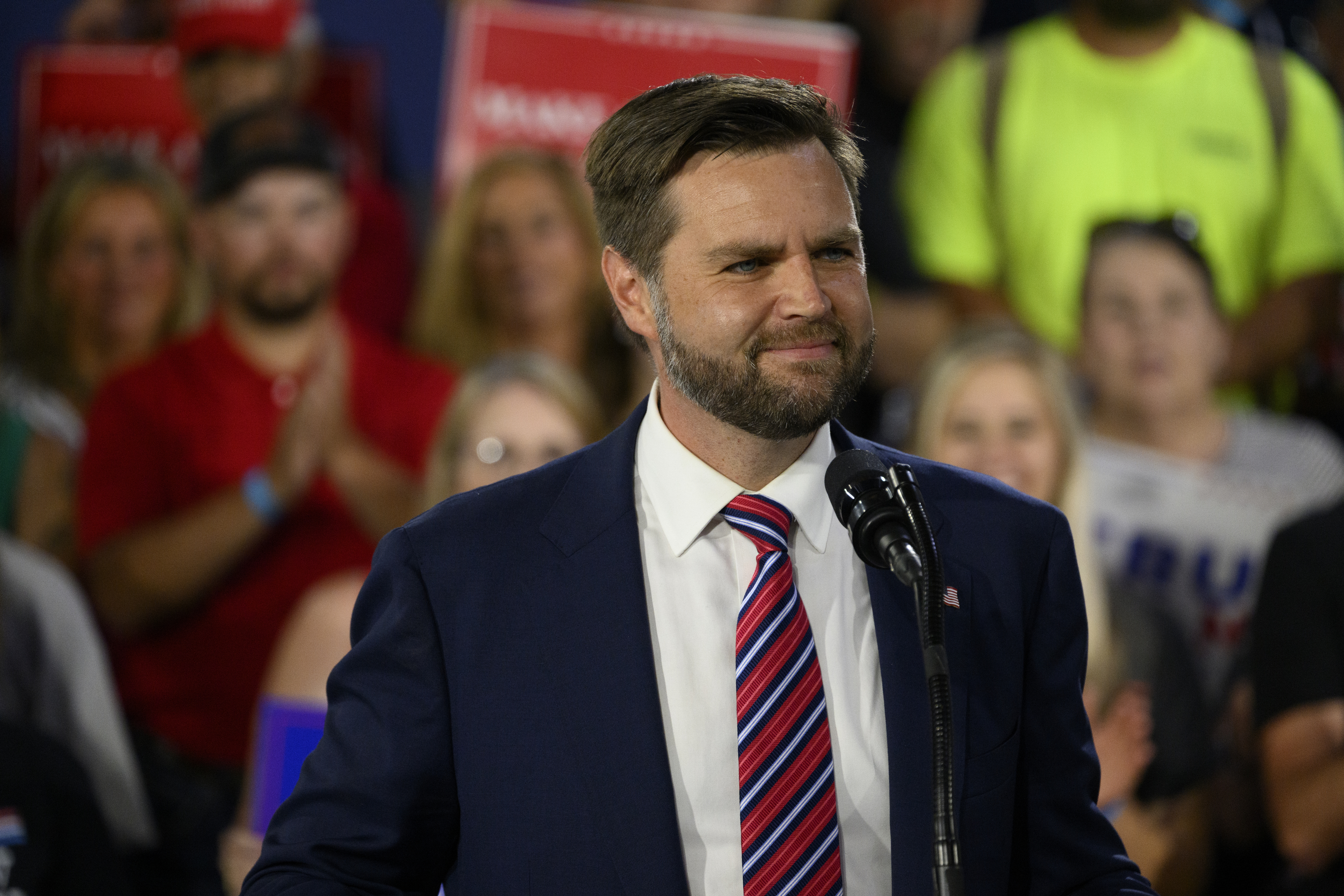 J.D. Vance stands at a podium speaking to a crowd, wearing a suit and striped tie. Fans and supporters are visible behind him