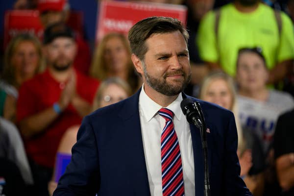 J.D. Vance stands at a podium speaking to a crowd, wearing a suit and striped tie. Fans and supporters are visible behind him