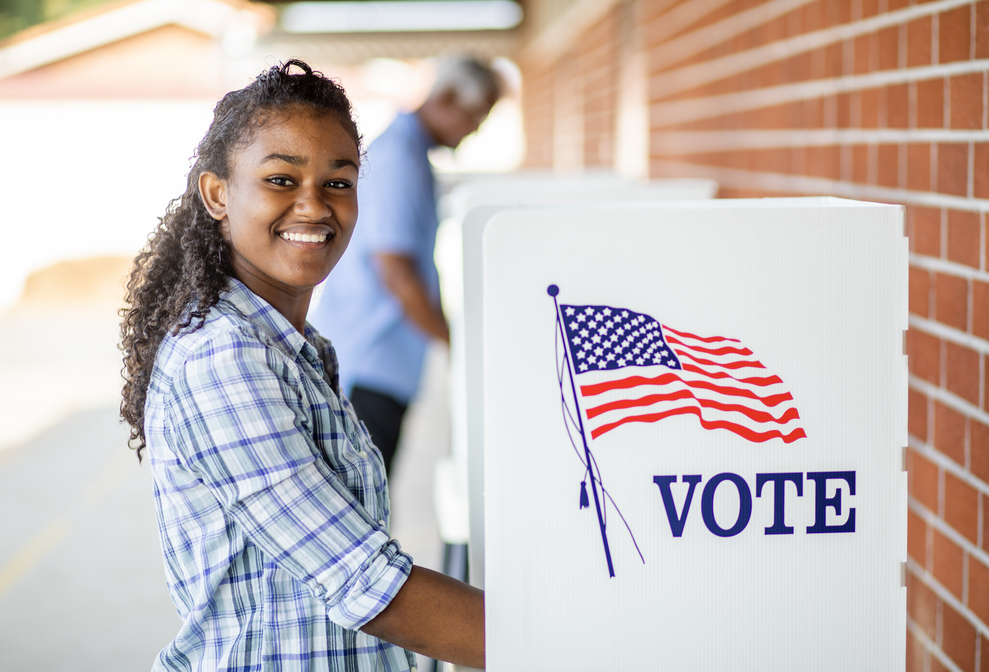 Woman smiling at a voting booth with the word &quot;VOTE&quot; and a U.S. flag on it. Another person is seen in the background at a different booth