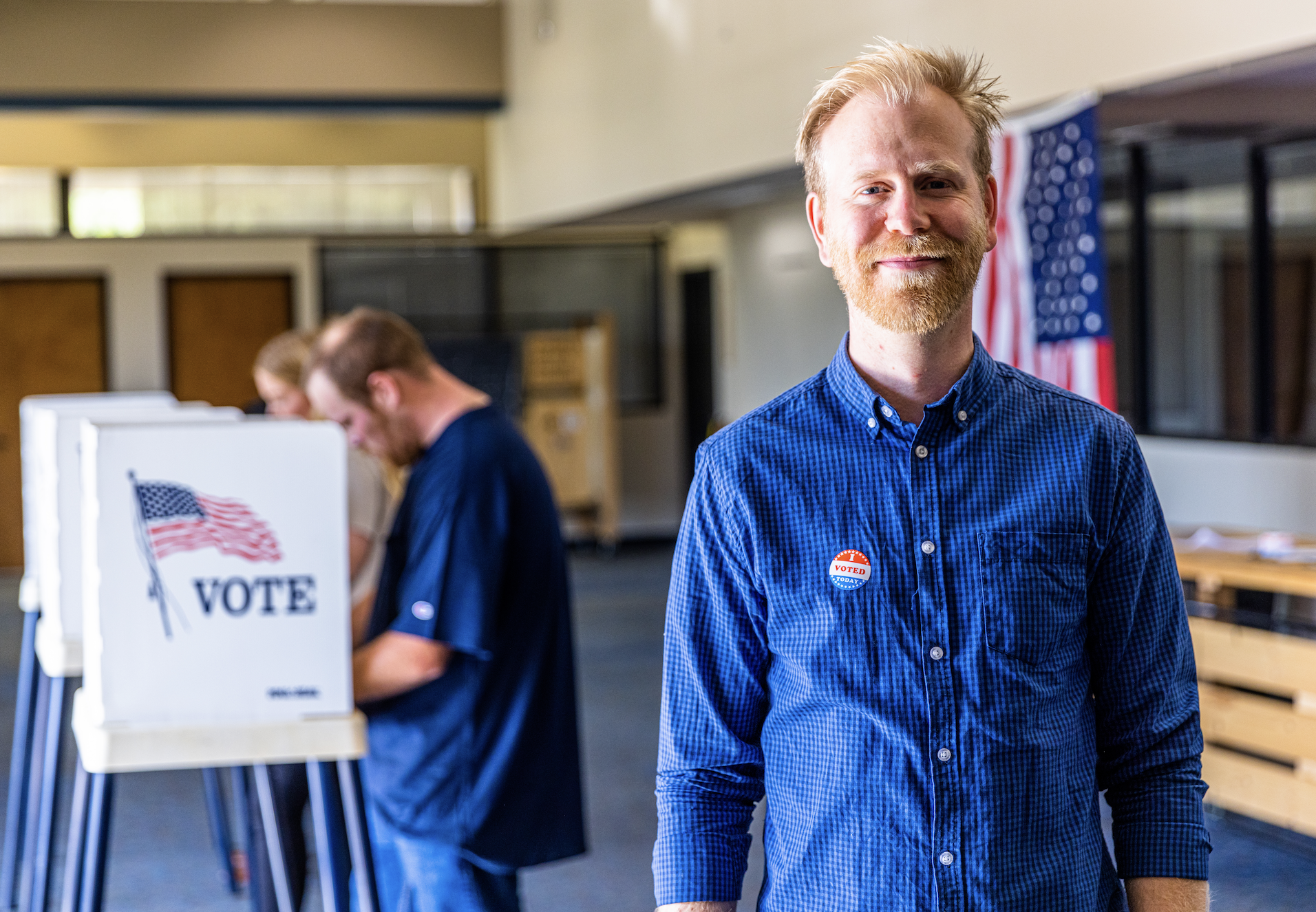A man stands smiling near voting booths in a polling station. Other people are seen casting their votes in the background