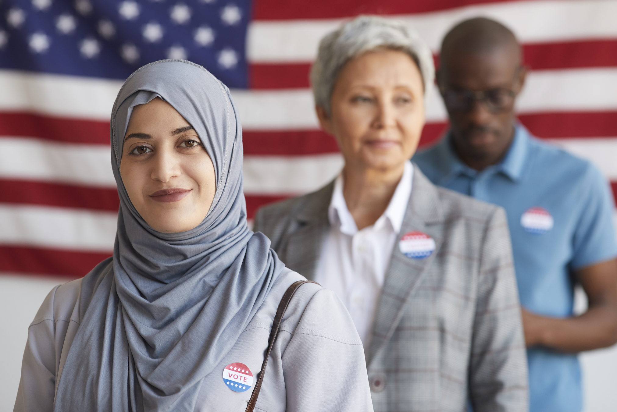 Three people standing in front of a U.S. flag wearing &quot;I Voted&quot; stickers, with one woman in a hijab in the foreground and two others in the background