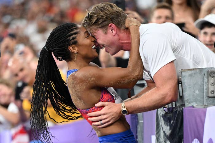 Tara in sportswear being held by Hunter leaning over from the stands, both smiling with their faces pressed together